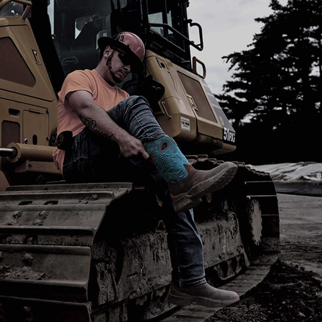 A man pulling on Phantom Rider Stinger Boots while sitting on the treads of a bulldozer.