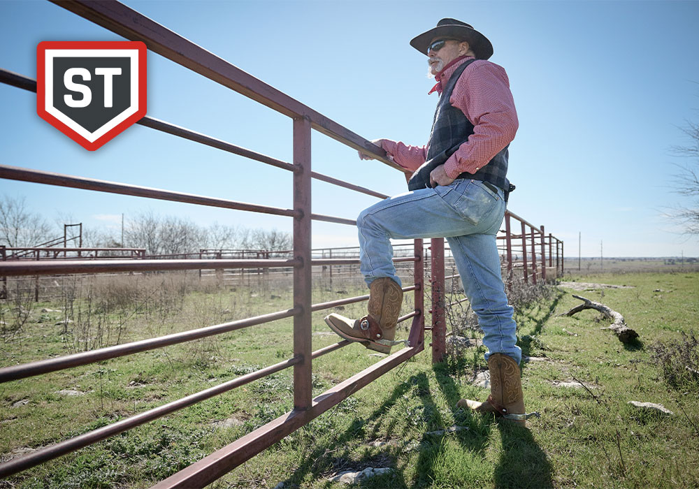 A cowboy leaning over a fence wearing the Dwight Steel toe western boot in brown. ST (Steel Toe) feature badge.