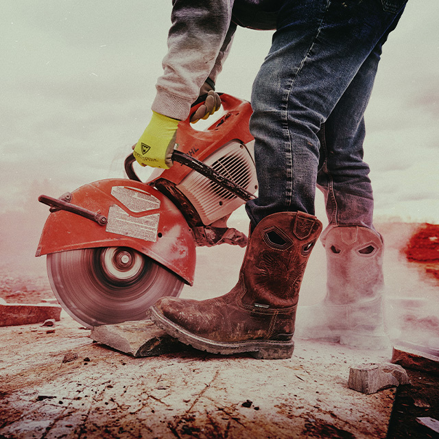 A person wearing the Dustie Comp Toe western boot in brown and using a circular saw on a log.