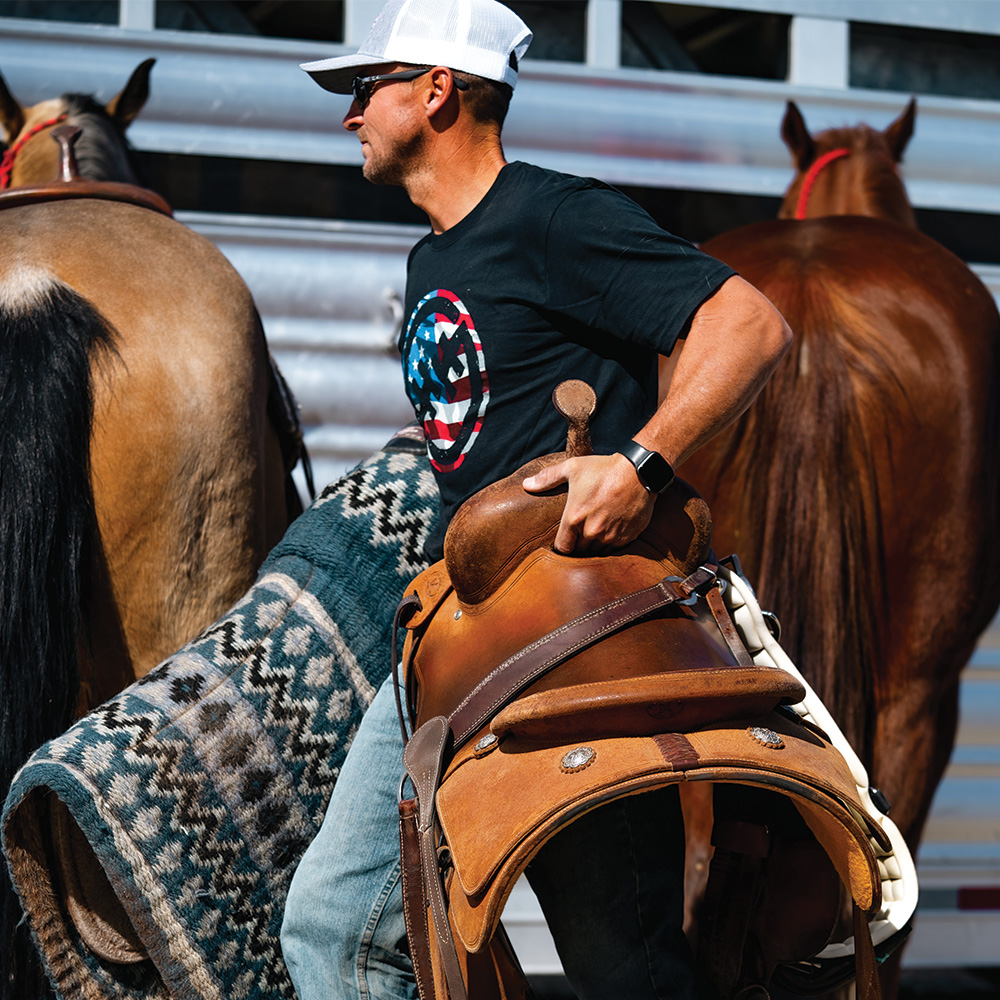 A man carrying horsing gear and walking away from their horses.