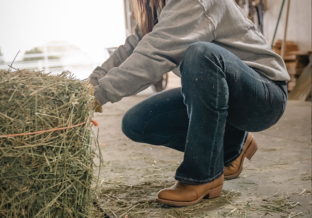 A woman wearing a Double-H boot in brown and wrapping a rope around hay.